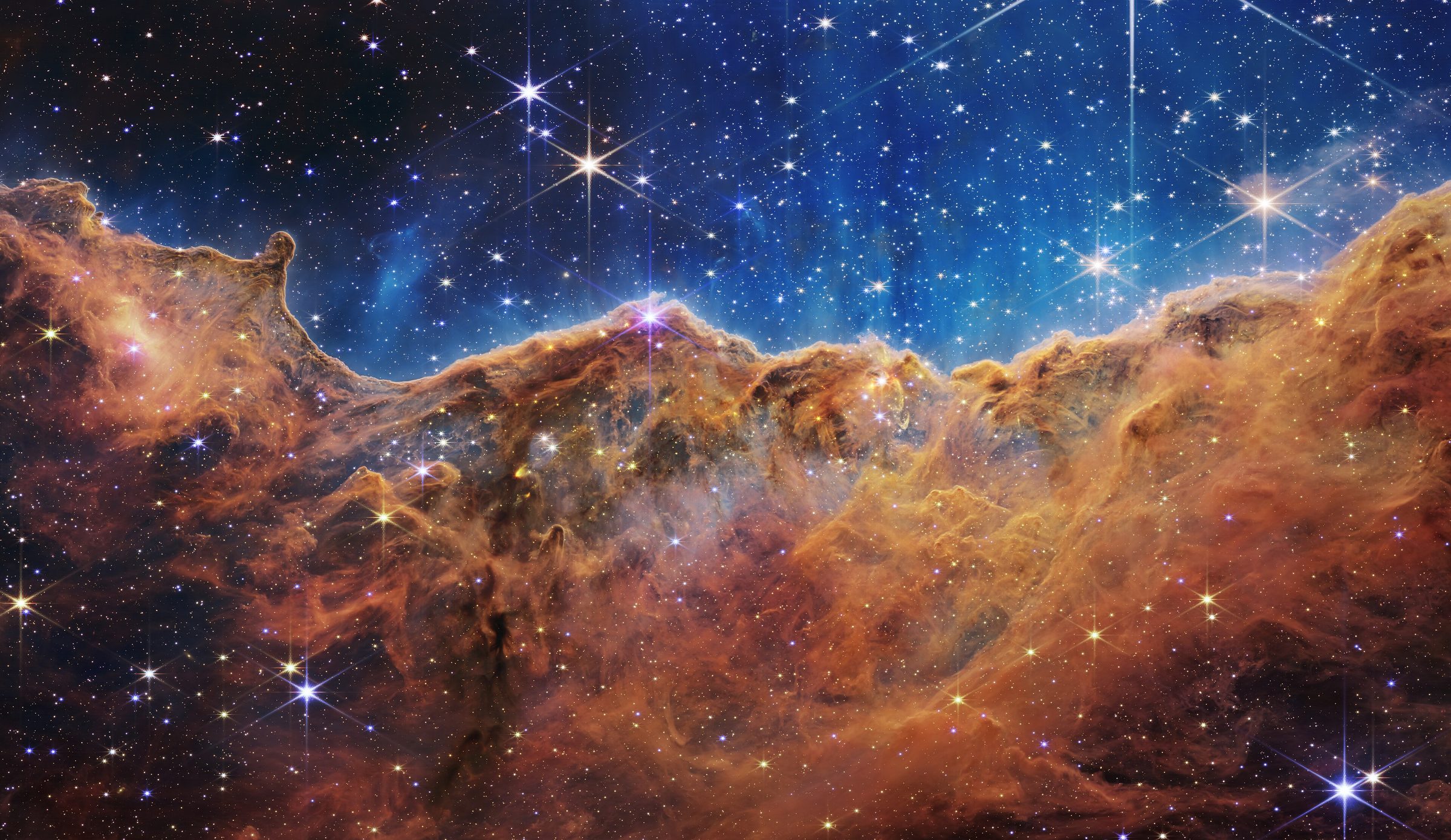 The Cosmic Cliffs of the Carina Nebula seen by the James Webb Space Telescope: a ridge of orange gas and dust under a blue sky thick with young stars.